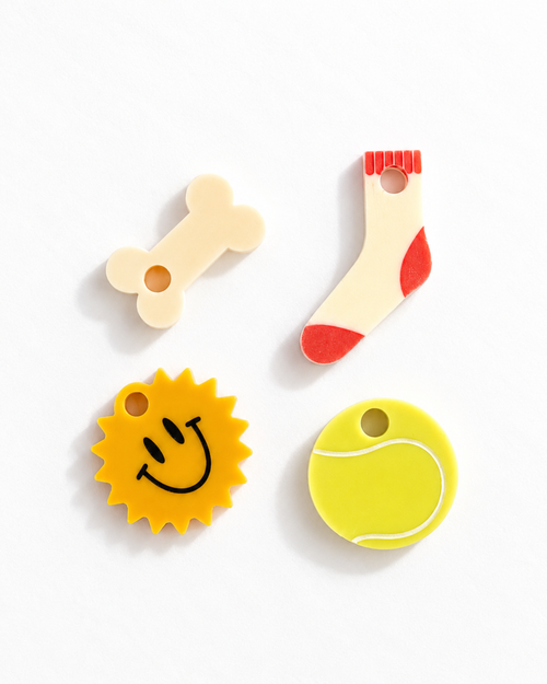 Four colorful dog toy shapes on a white background: a cream bone, a cream sock with red accents, a yellow sun with a black smiley face, and a yellow tennis ball.